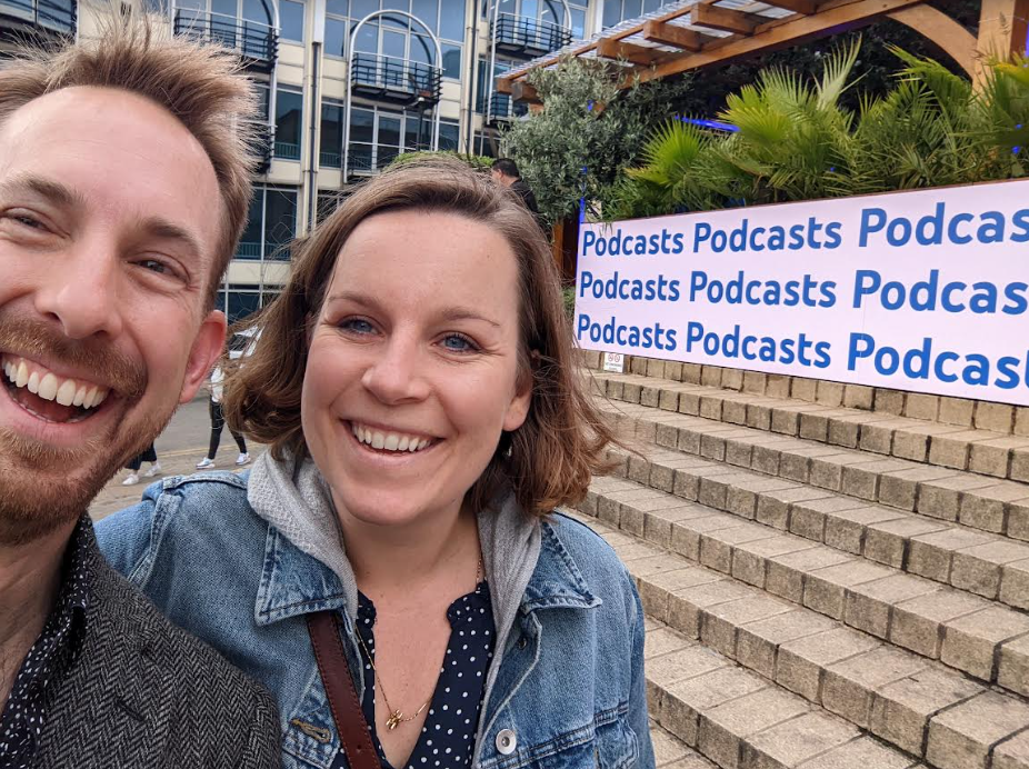 Izabela and Mike Russell of Music Radio Collective outside The Podcast Show London event in May 2022. Behind them is a poster saying "podcasts" nine times.